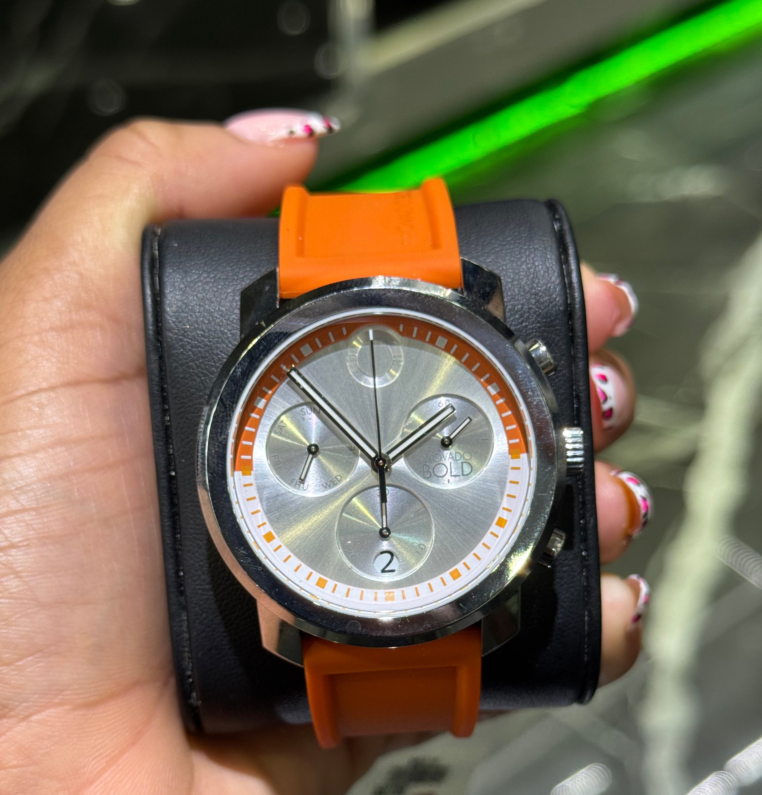 Hand holding a watch with an orange strap against a blurred background