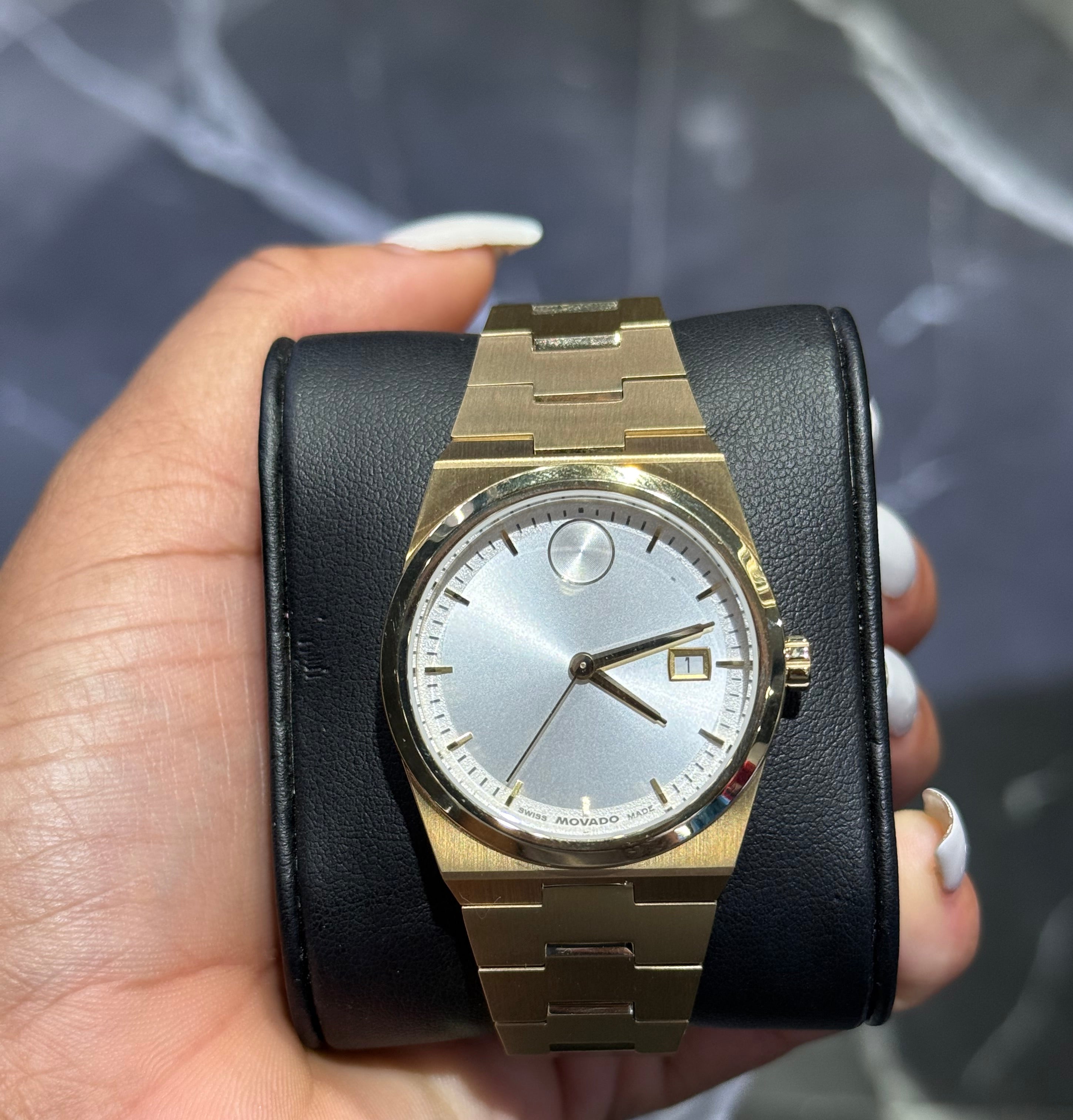 Gold wristwatch with black leather strap held in a hand against a blurred background