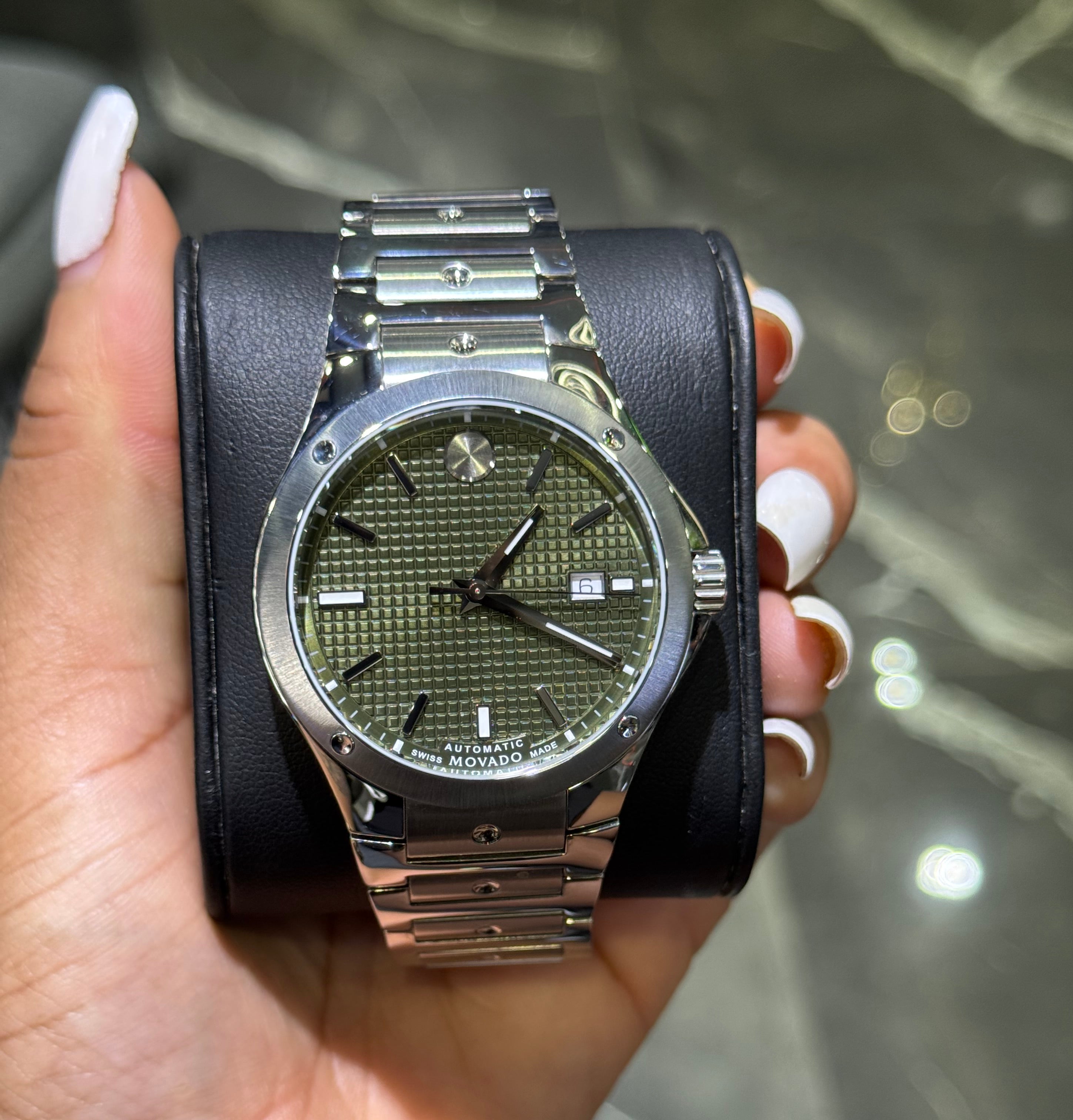 Hand holding a watch with a black strap and green face against a blurred background