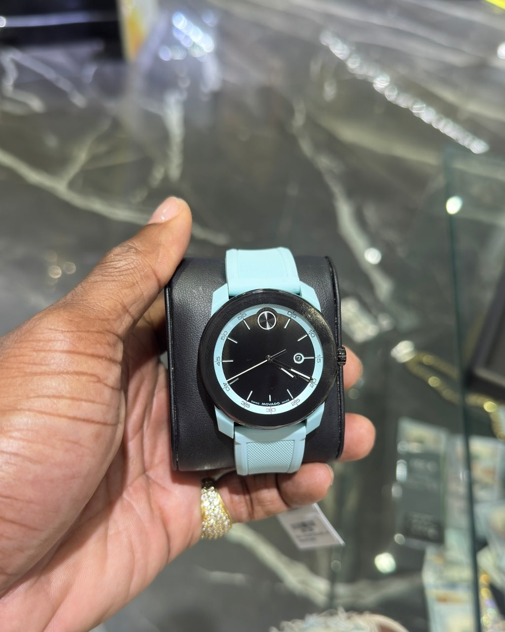 Hand holding a watch with a black face and light blue strap against a reflective surface.