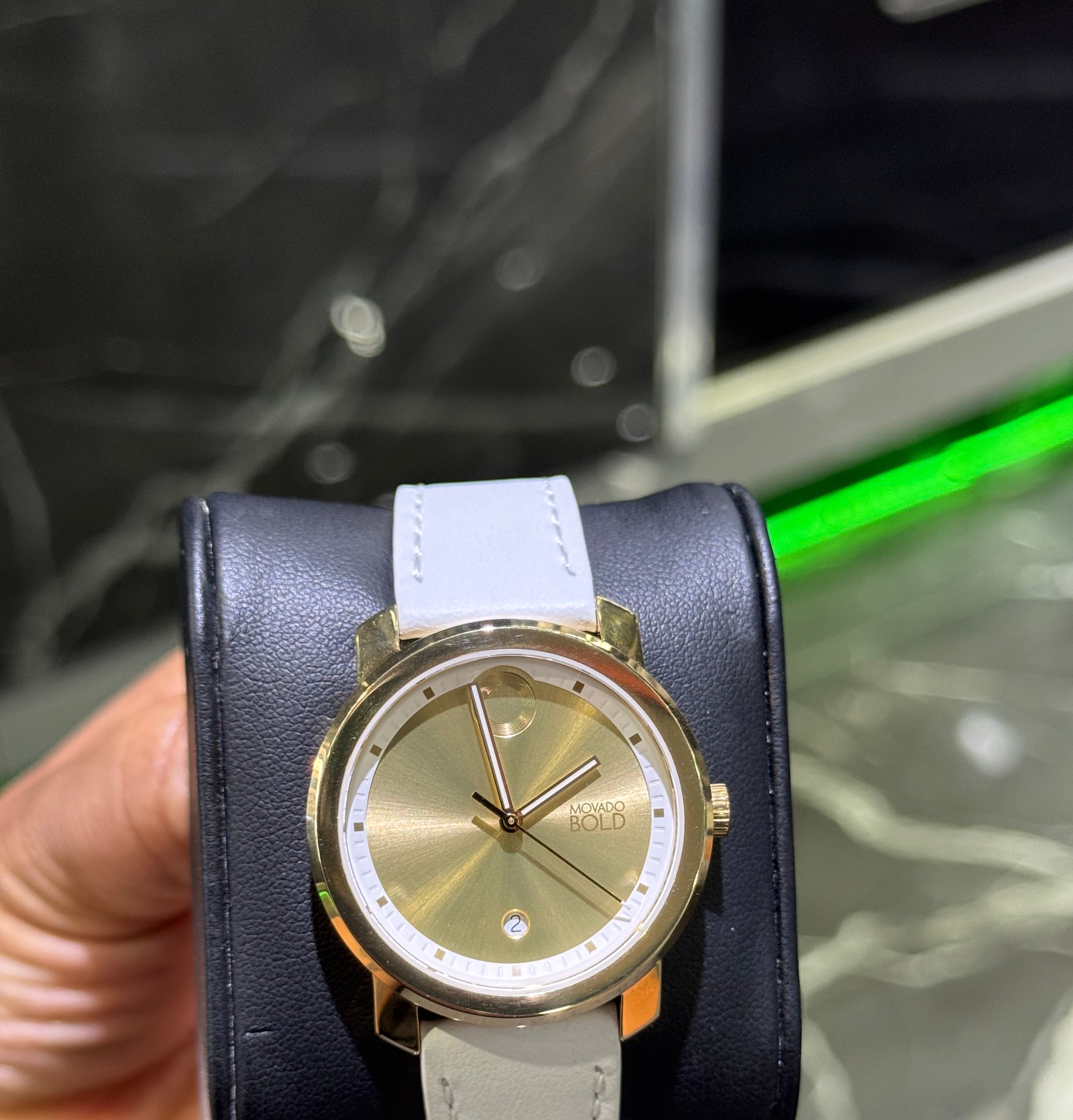 Gold watch with a white strap held by a hand against a blurred background