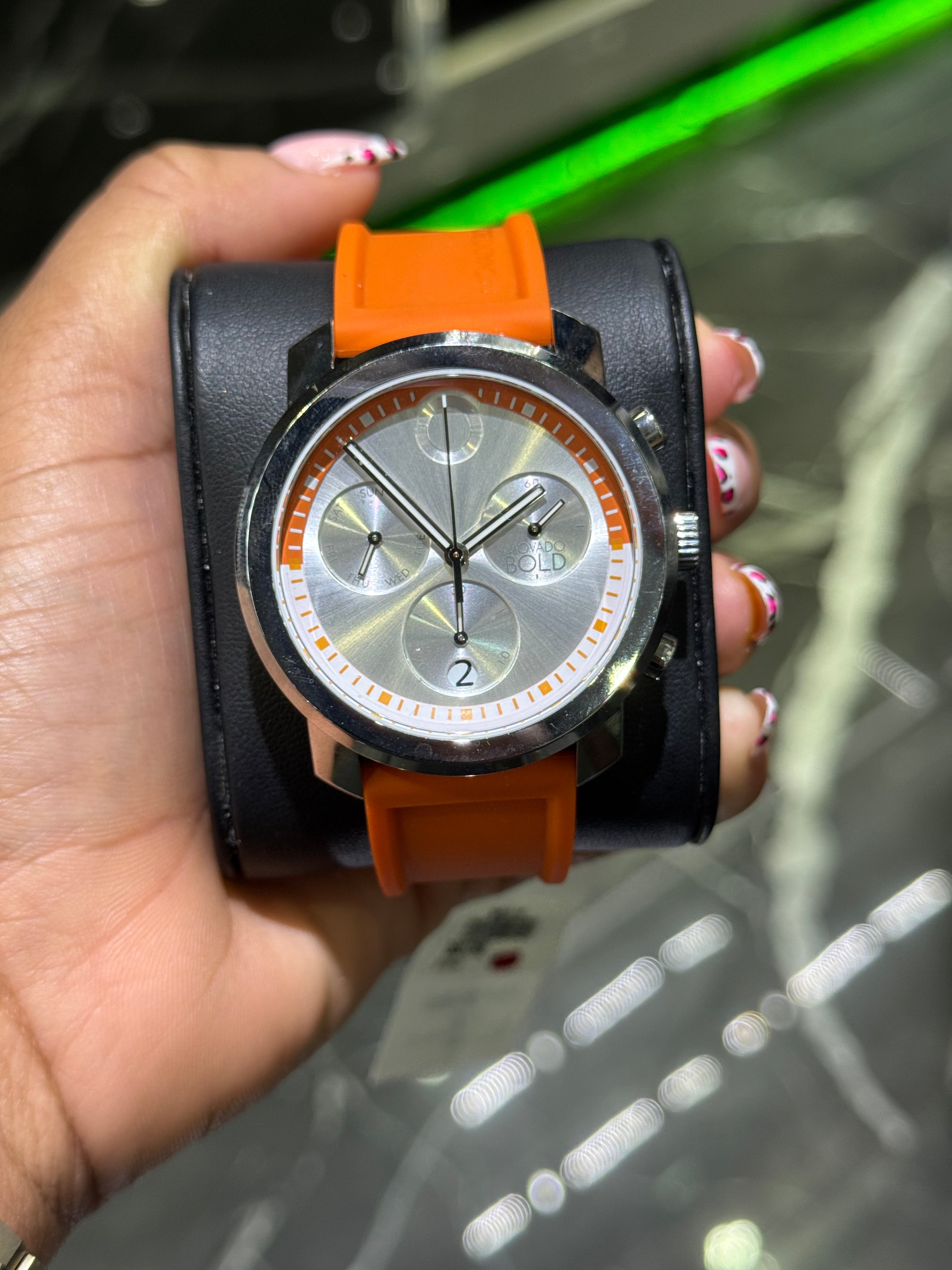 Hand holding a watch with an orange strap against a blurred background