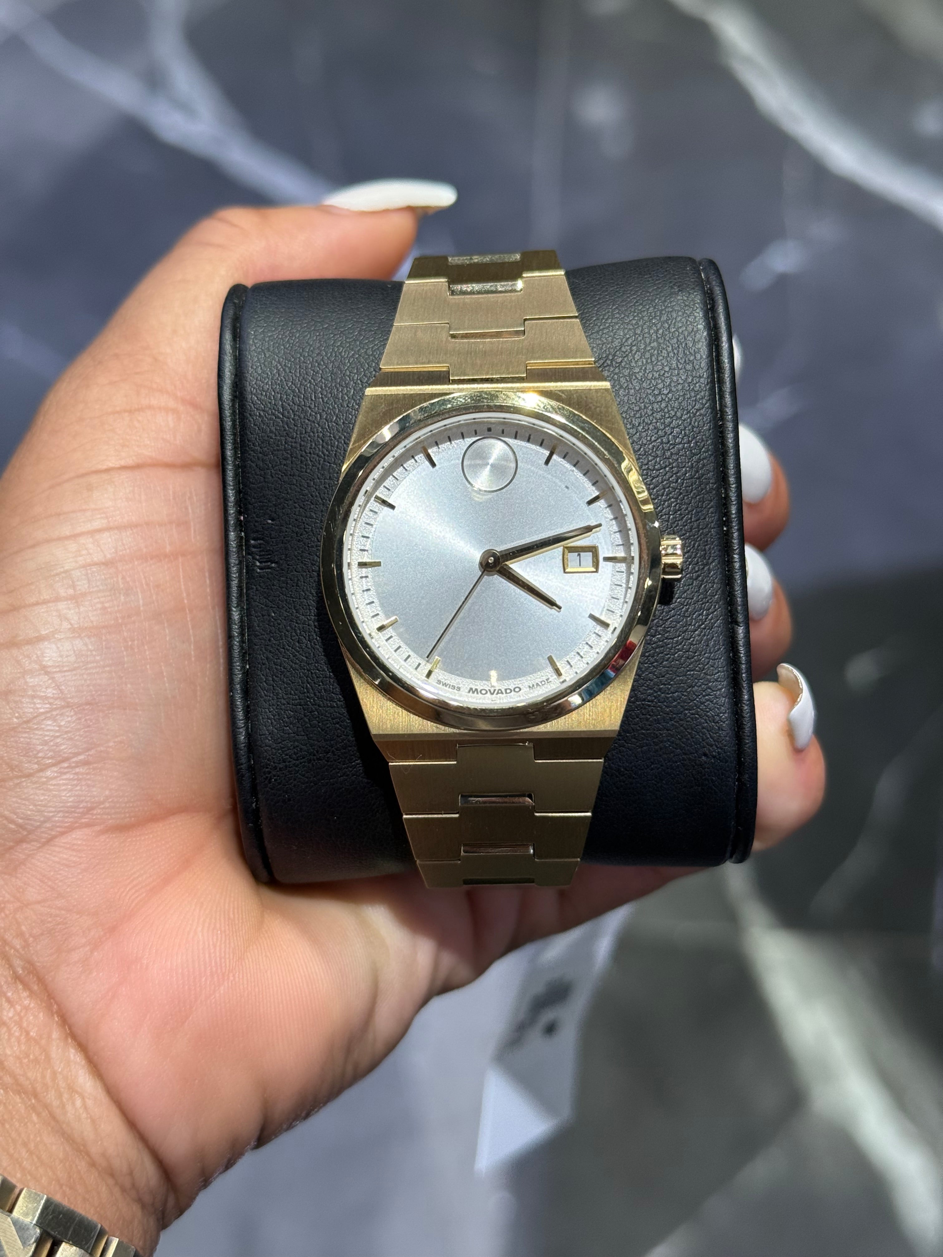 Gold wristwatch with black leather strap held in a hand against a blurred background