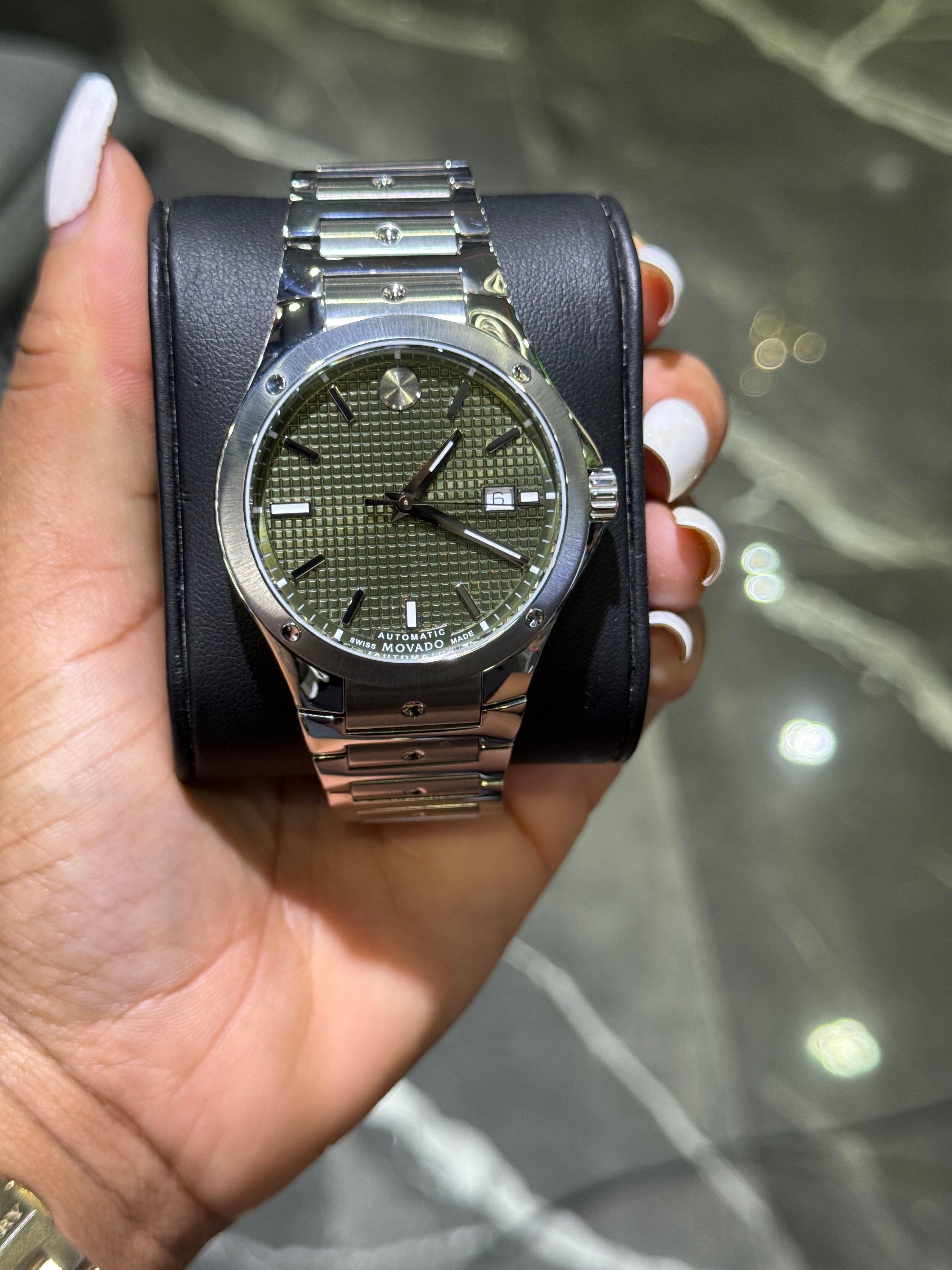 Hand holding a watch with a black strap and green face against a blurred background