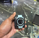 Hand holding a watch with a black face and light blue strap against a reflective surface.