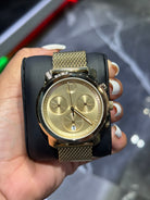 Gold watch with black strap held in a hand against a blurred background