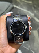Hand holding a silver watch with a black face against a blurred background